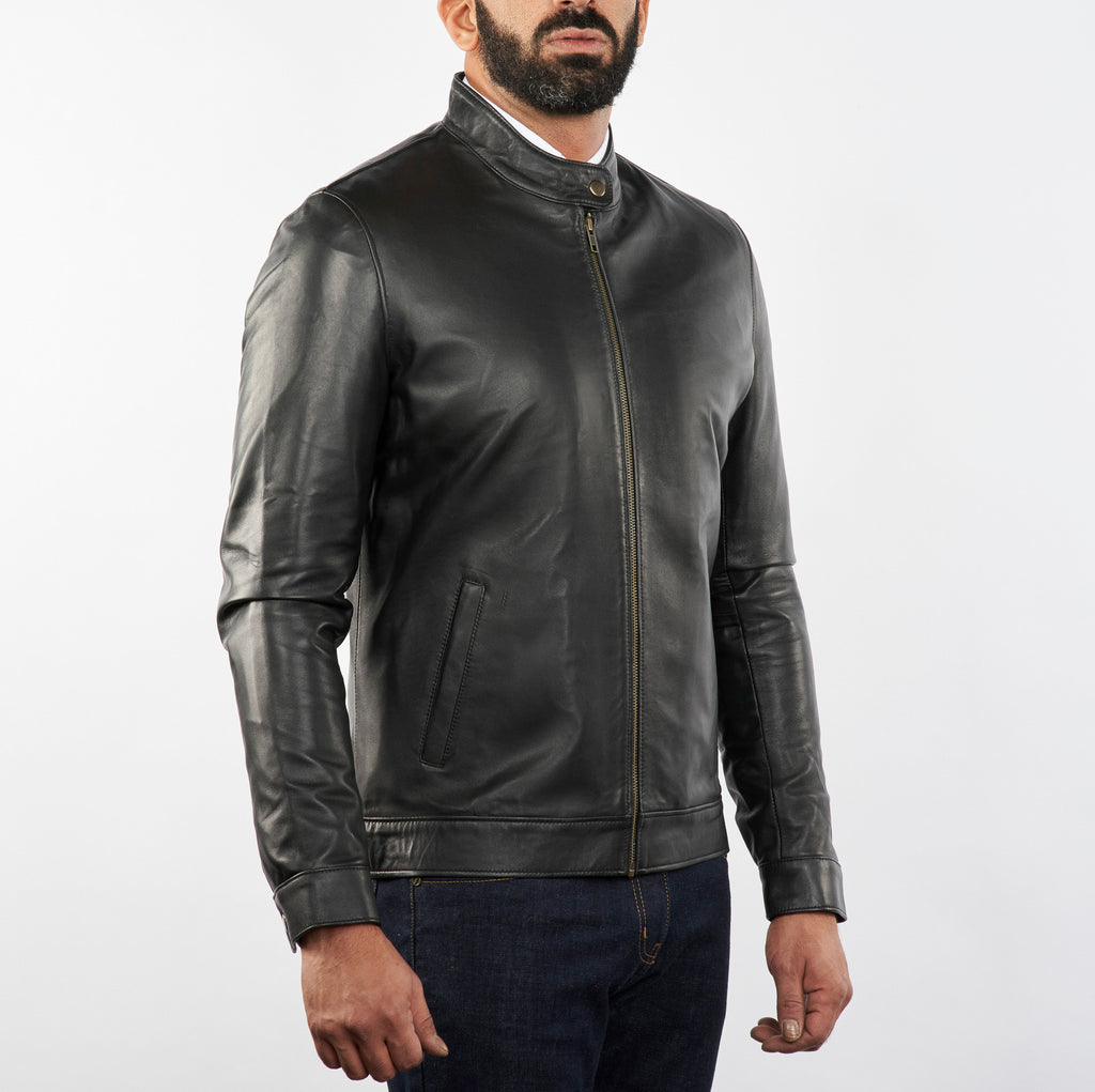 A man wearing a black leather bomber jacket with a stand-up collar and front zipper, paired with dark blue jeans, photographed against a plain light gray studio background.