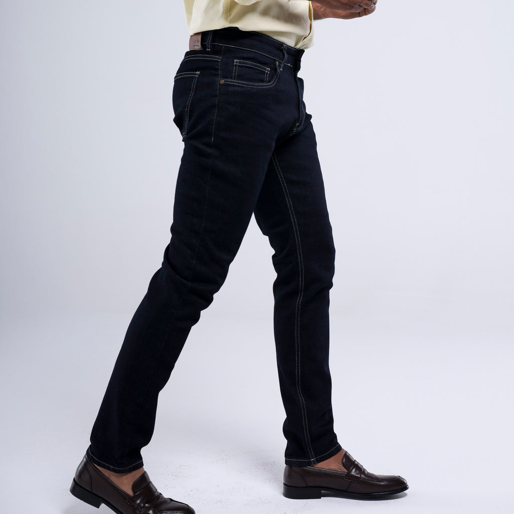 A person wearing slim-fit black jeans with contrast white stitching, paired with a cream shirt and dark brown leather loafers, shown from the waist down against a light gray studio background.