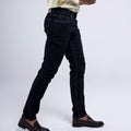 A person wearing slim-fit black jeans with contrast white stitching, paired with a cream shirt and dark brown leather loafers, shown from the waist down against a light gray studio background.
