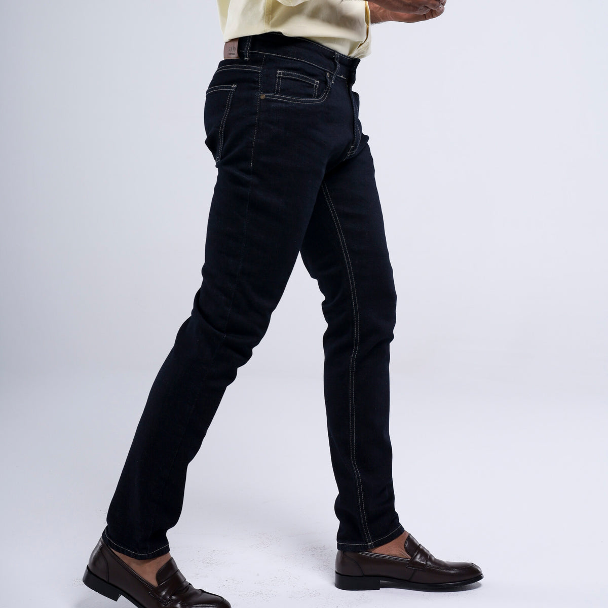A person wearing slim-fit black jeans with contrast white stitching, paired with a cream shirt and dark brown leather loafers, shown from the waist down against a light gray studio background.