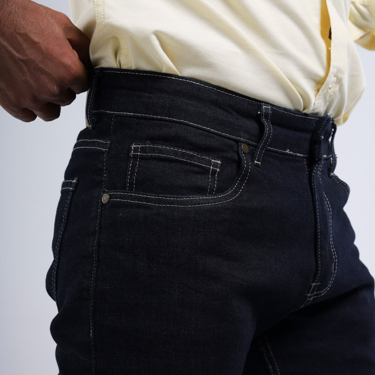 Close-up of a person wearing dark indigo denim jeans with white contrast stitching, pulling the waistband slightly with a hand, wearing a cream-colored collared shirt.