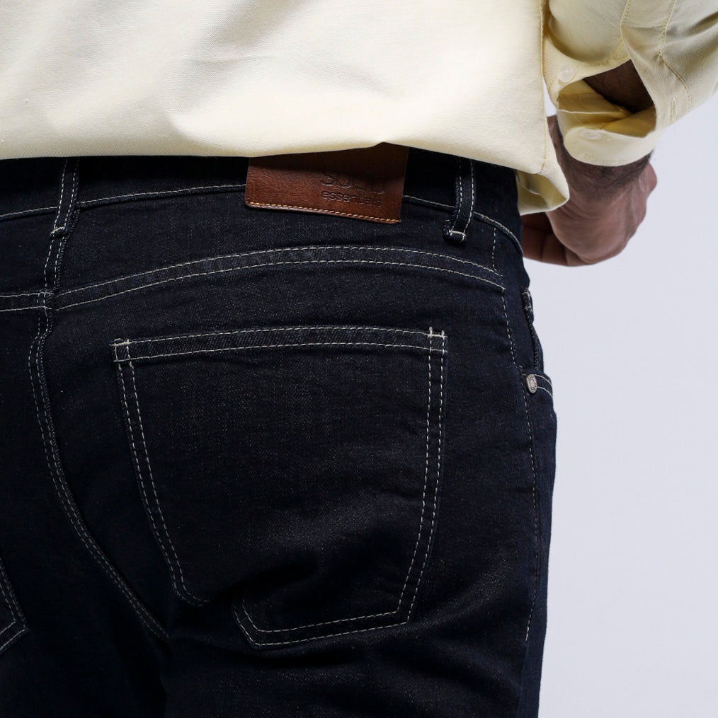 Close-up back view of a person wearing dark indigo denim jeans with white contrast stitching and a brown leather patch at the waistband, paired with a light cream shirt.