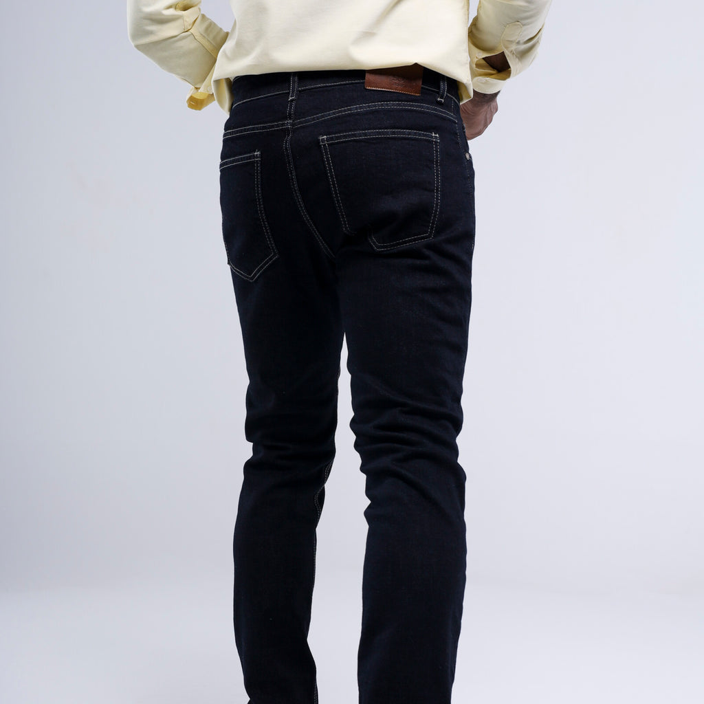 Rear view of a person wearing slim-fit dark indigo jeans with contrast stitching, paired with a cream-colored long-sleeve shirt, against a light gray studio background.