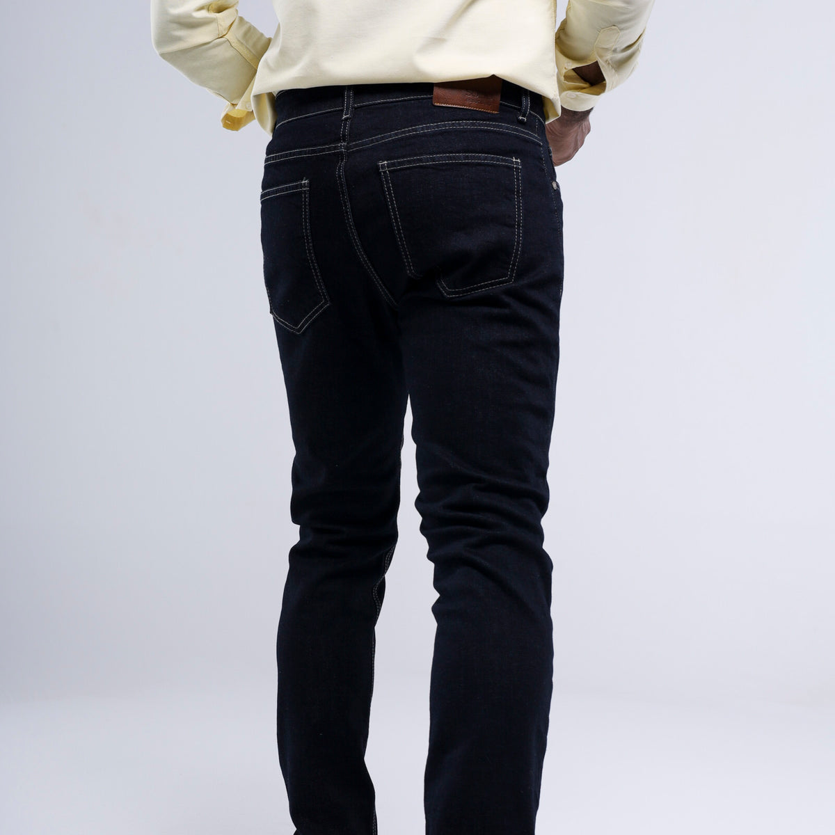 Rear view of a person wearing slim-fit dark indigo jeans with contrast stitching, paired with a cream-colored long-sleeve shirt, against a light gray studio background.