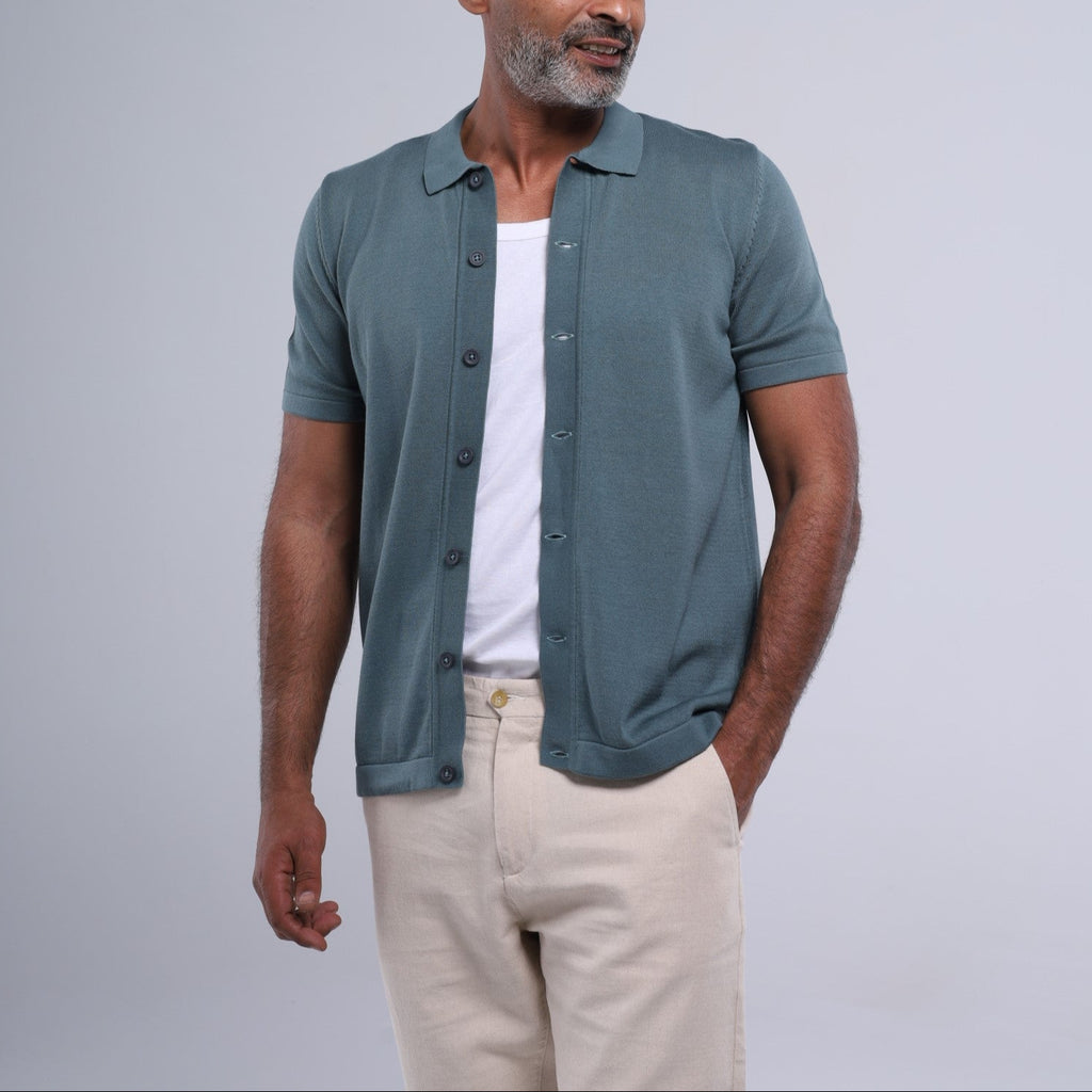 Man wearing a teal button-up shirt over a white t-shirt with beige pants on a light gray background