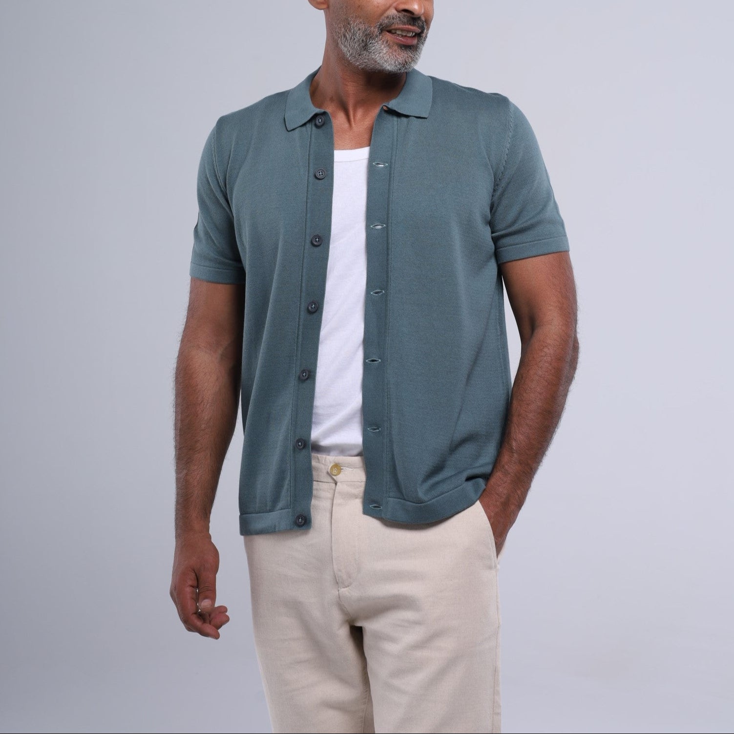 Man wearing a teal button-up shirt over a white t-shirt with beige pants on a light gray background