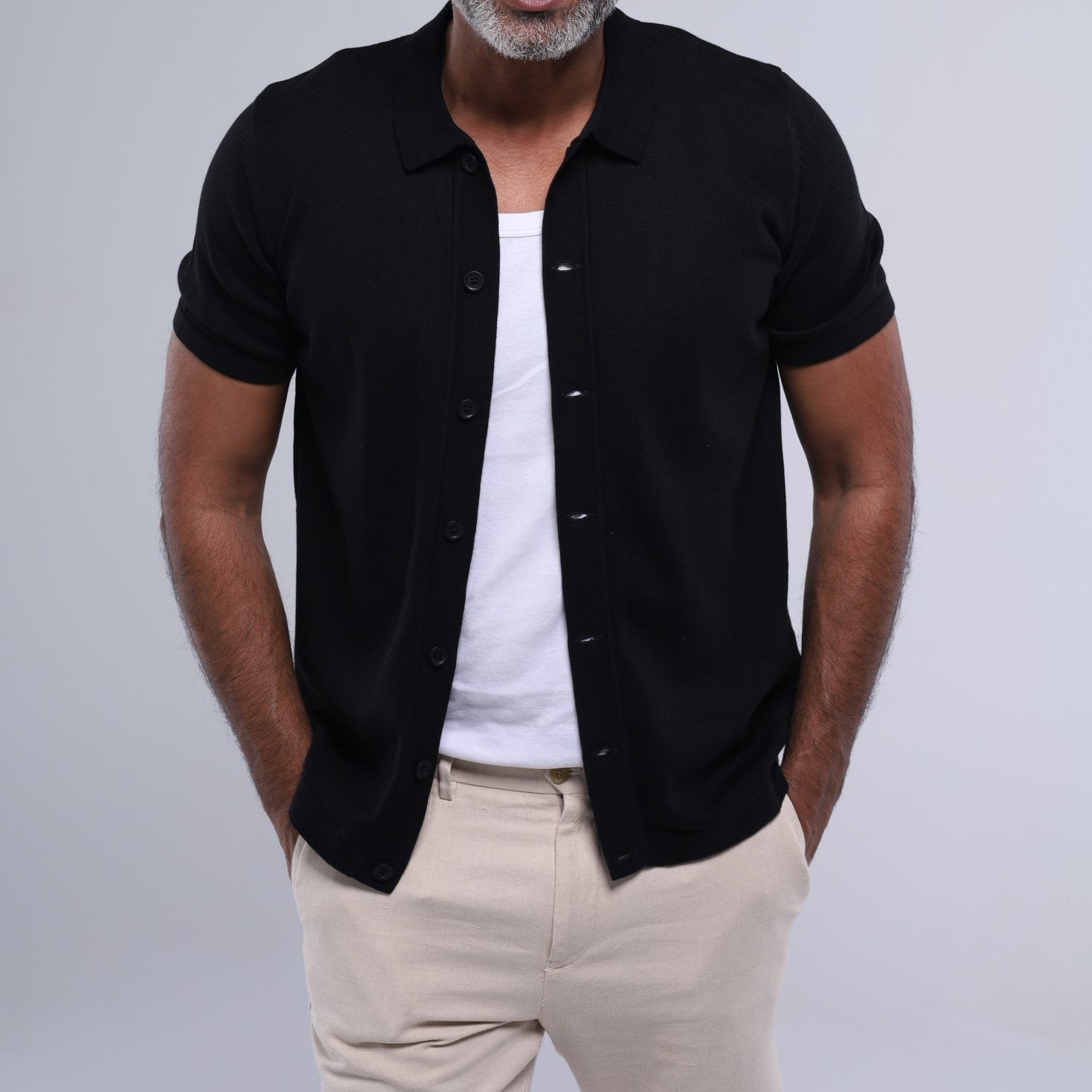 Man wearing a black button-up shirt over a white t-shirt with beige pants on a gray background