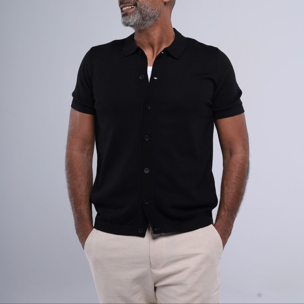 Man wearing a black short-sleeve button-up shirt and beige pants against a gray background