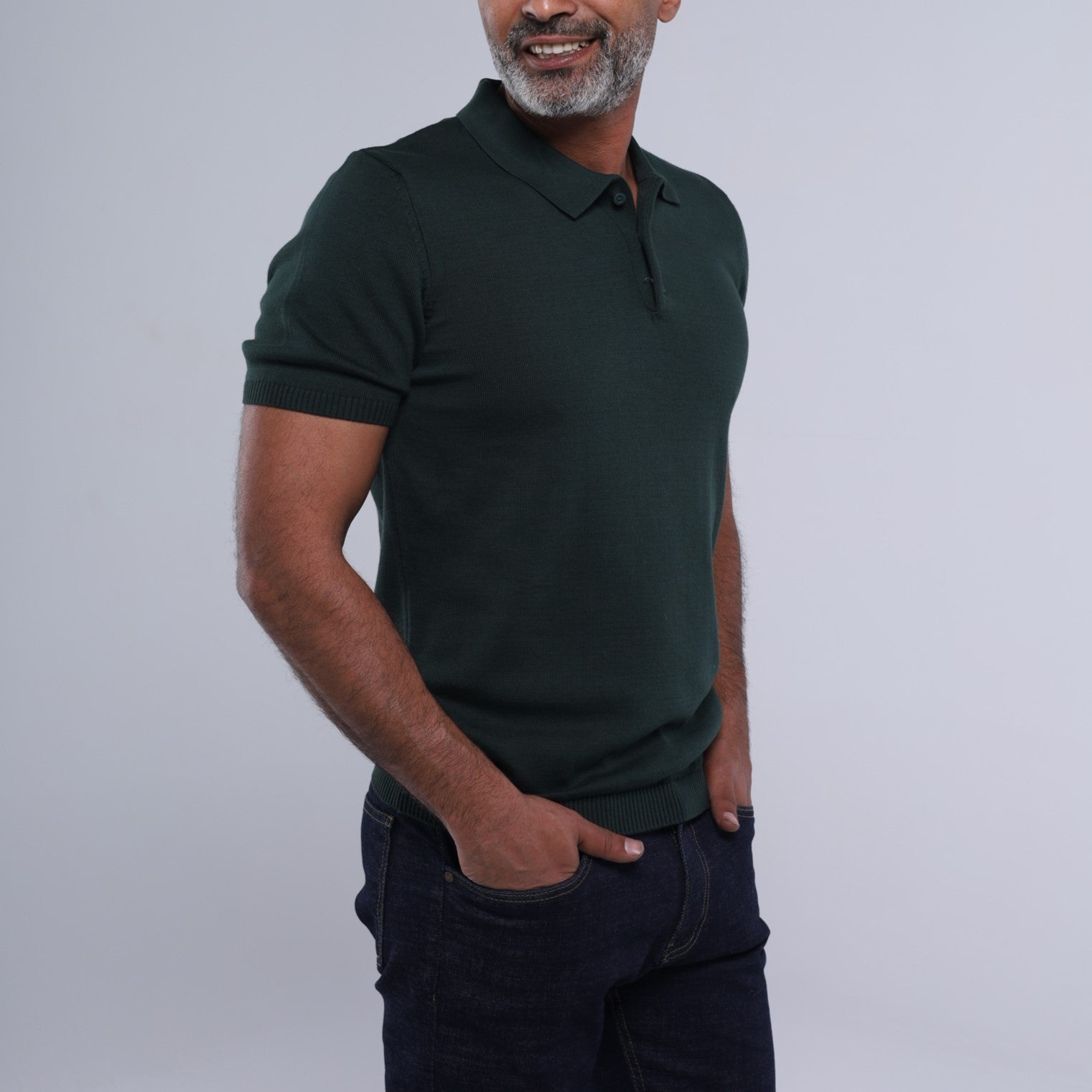 A man wearing a dark forest green short-sleeve polo shirt and dark blue jeans, standing against a light gray studio background.