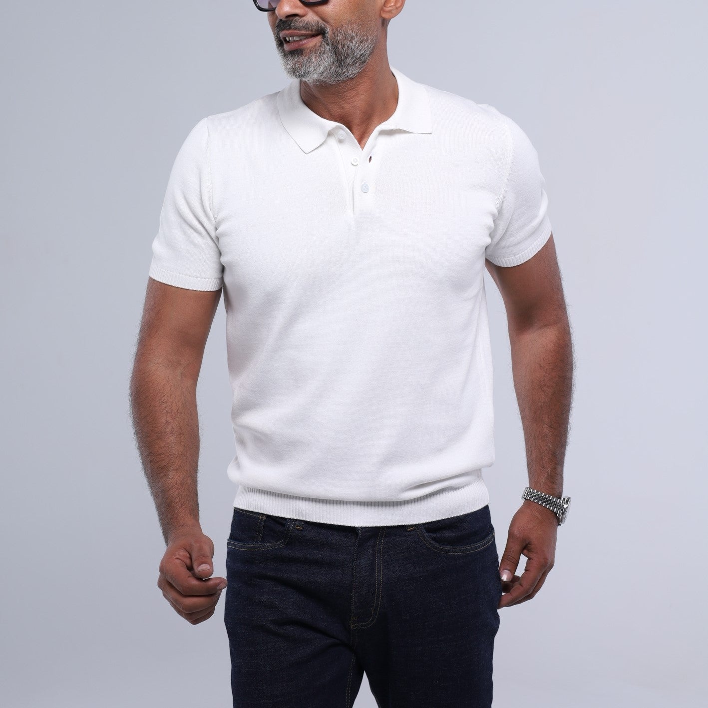 A man wearing a white short-sleeve knit polo shirt and dark blue jeans, standing against a light gray studio background.