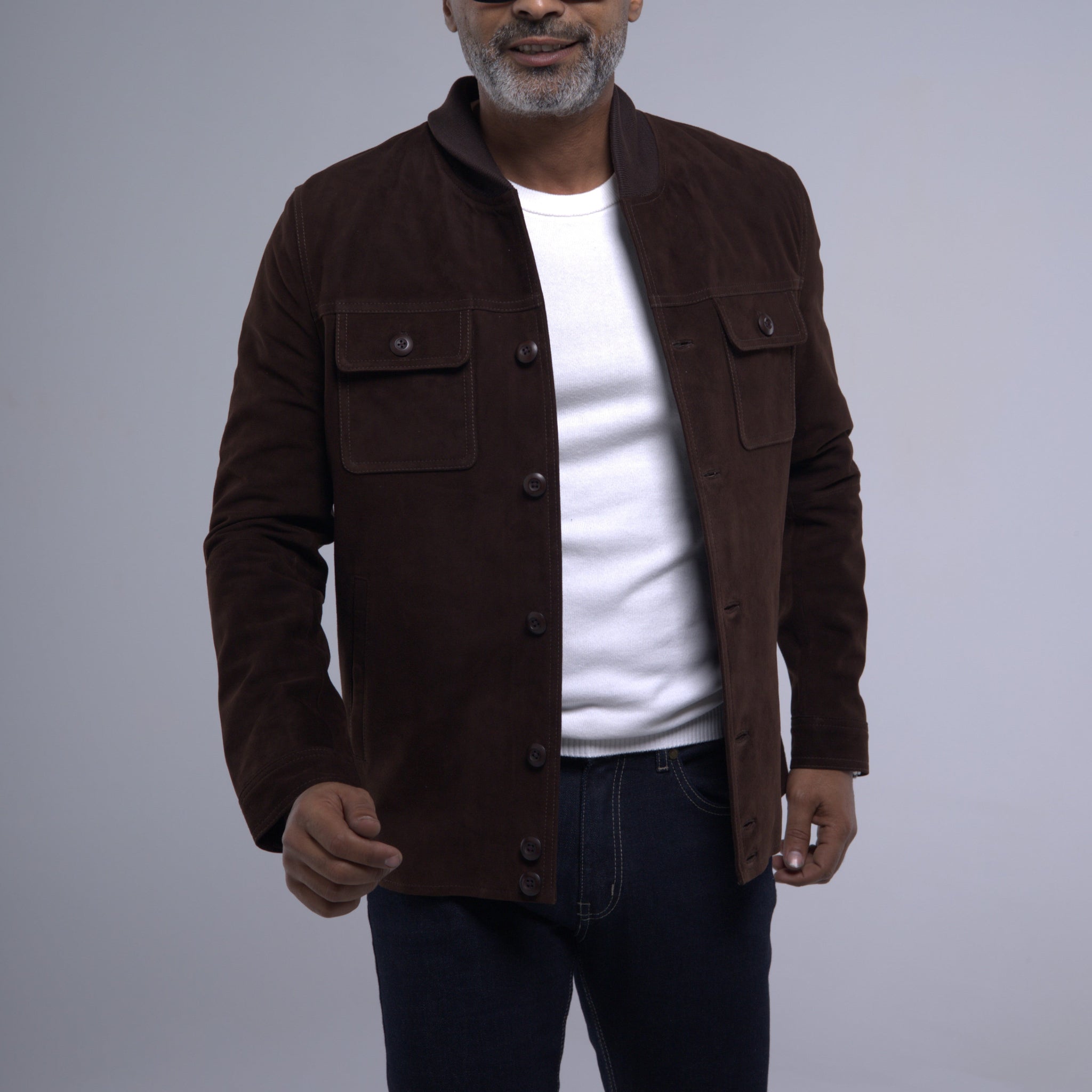 A man wearing a dark brown suede jacket over a white t-shirt and dark jeans, standing against a neutral gray studio background.