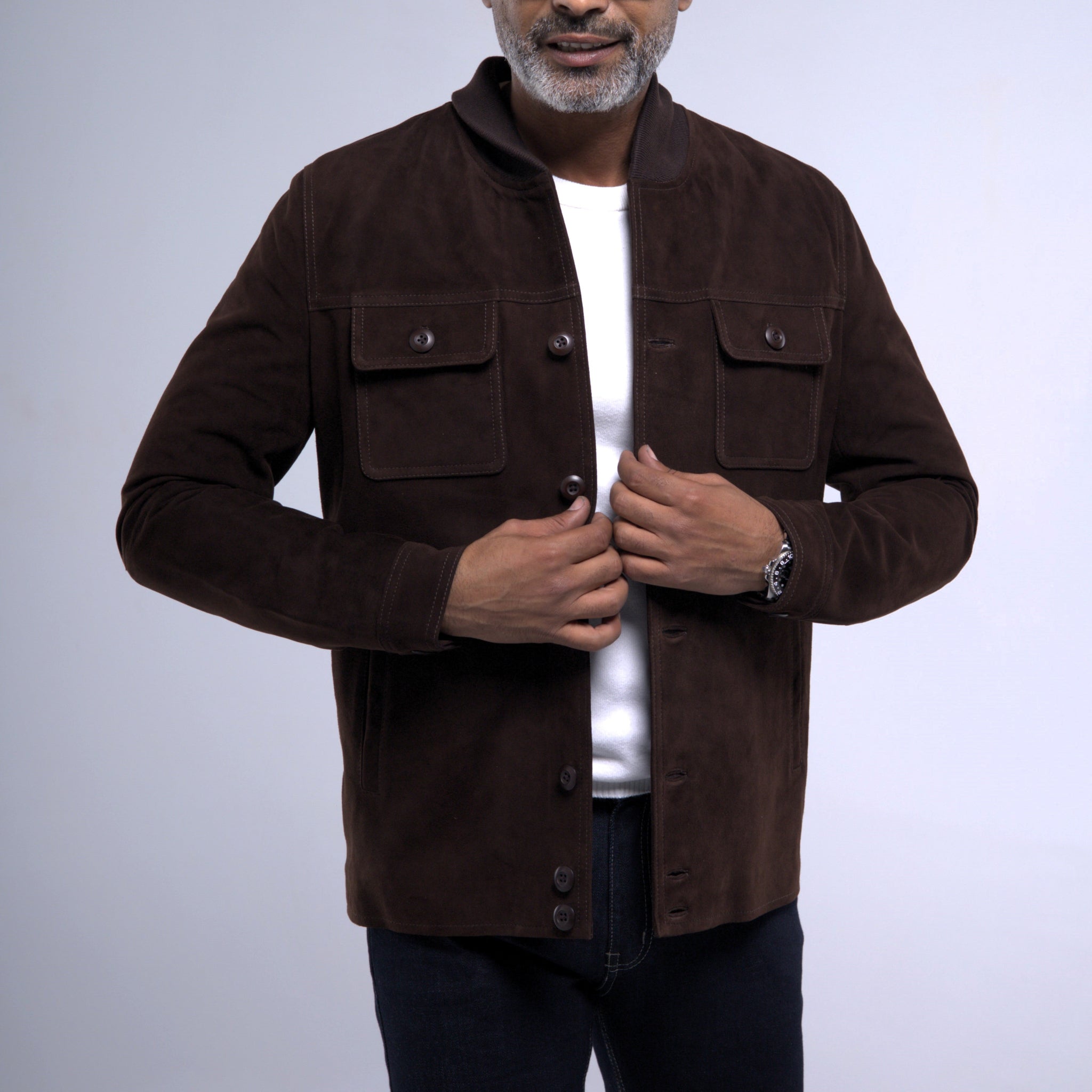 A man wearing a dark brown suede jacket over a white t-shirt and dark jeans, shown from the chin down against a light gray studio background.