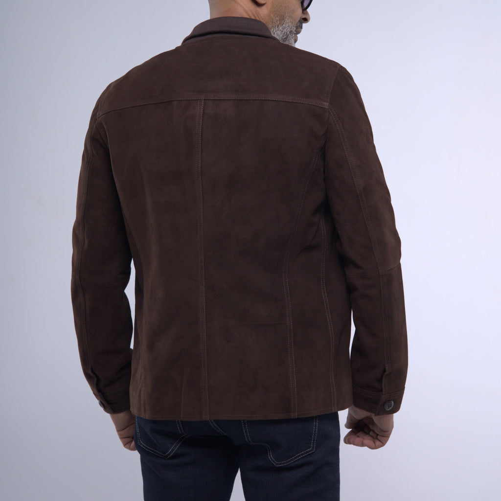 A man viewed from the back wearing a dark brown suede jacket and dark blue jeans against a light gray studio background.