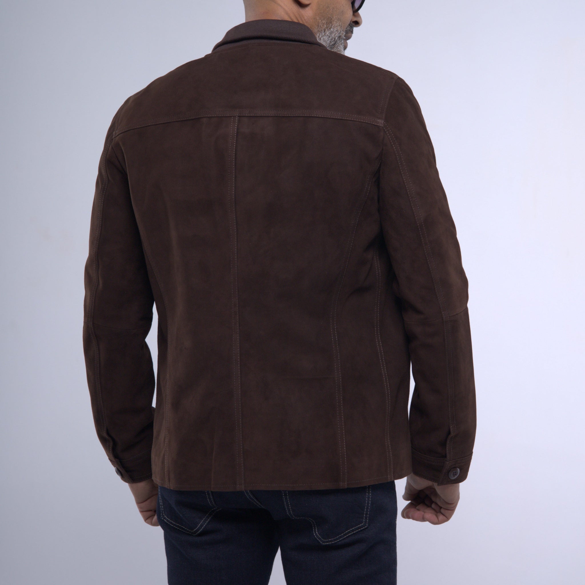 A man viewed from the back wearing a dark brown suede jacket and dark blue jeans against a light gray studio background.