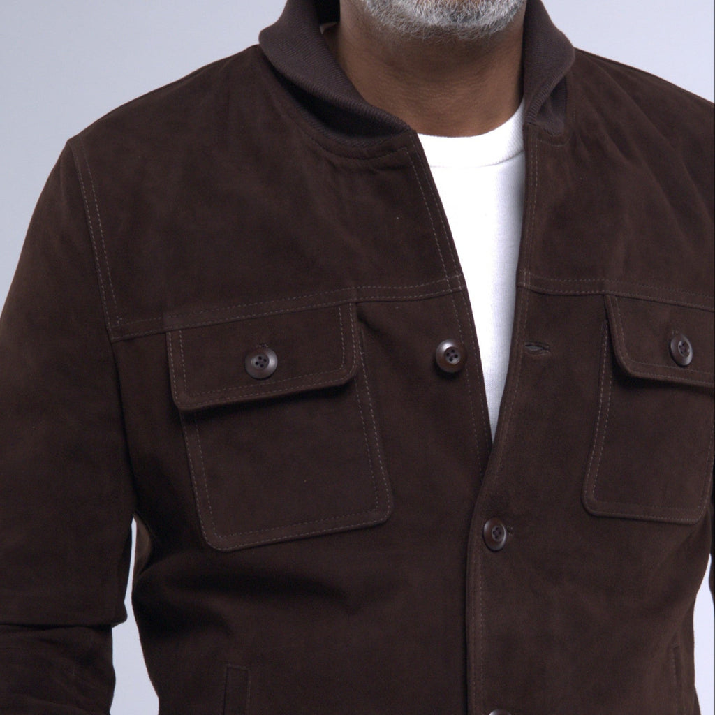 A man wearing a dark brown suede jacket over a white t-shirt and dark jeans, photographed against a light gray studio background.