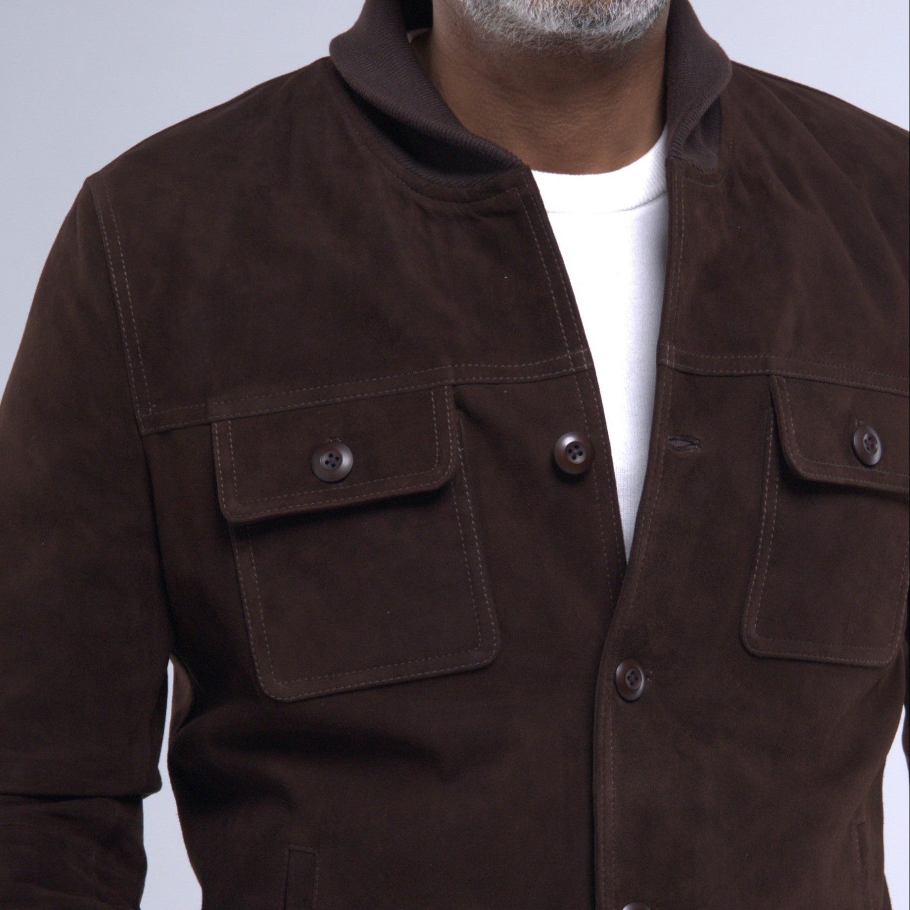 A man wearing a dark brown suede jacket over a white t-shirt and dark jeans, photographed against a light gray studio background.