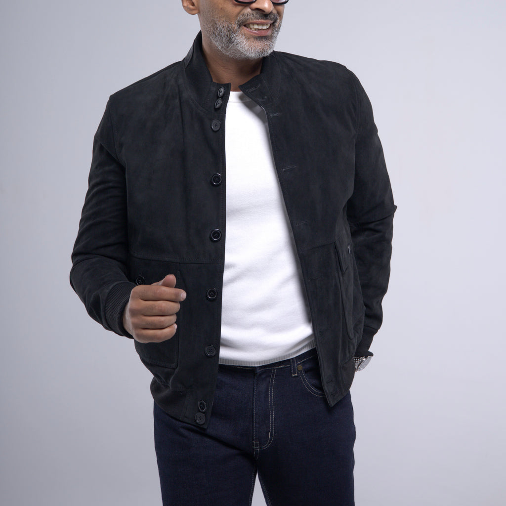 A man wearing a black suede jacket over a white t-shirt and dark jeans, standing against a light gray studio background.