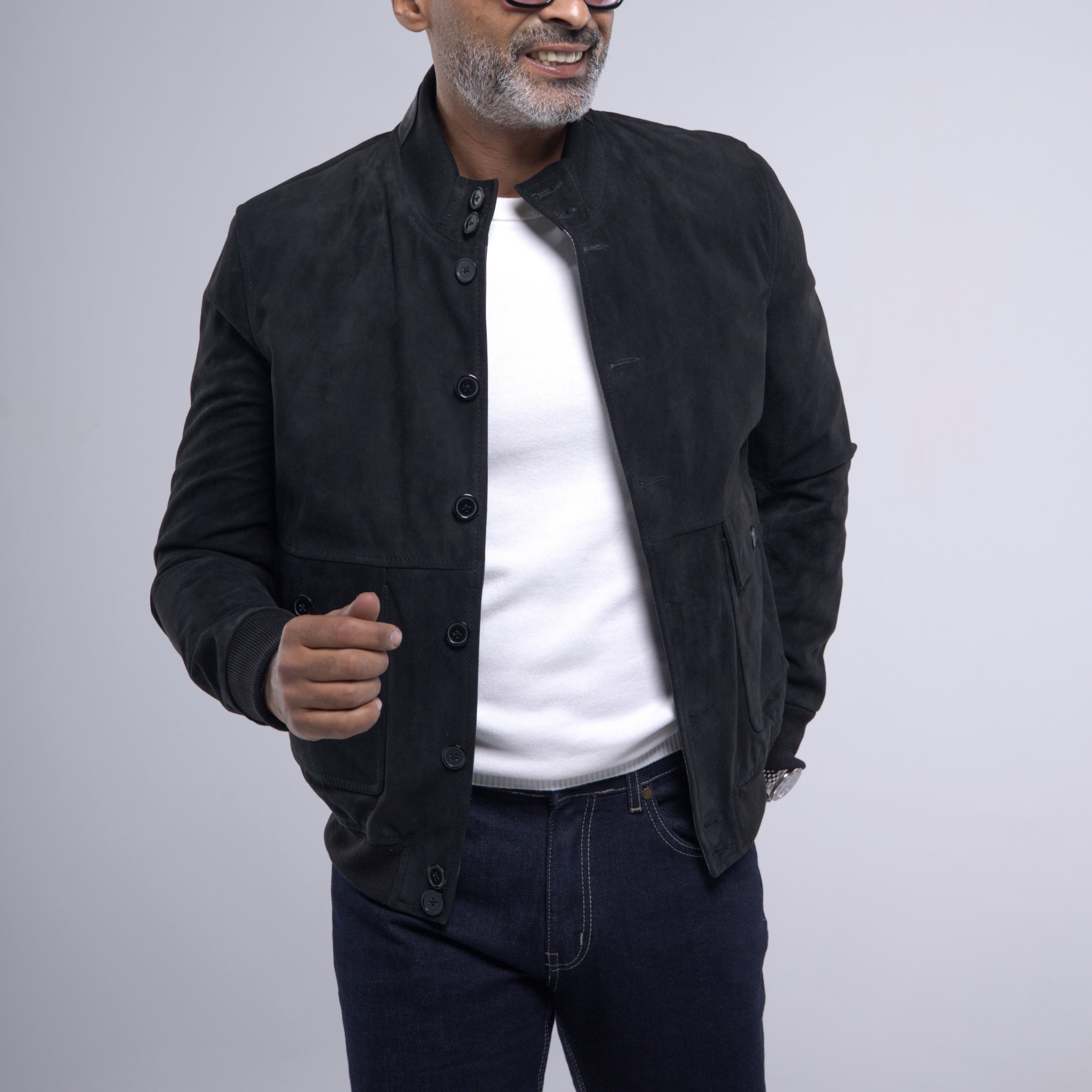 A man wearing a black suede jacket over a white t-shirt and dark jeans, standing against a light gray studio background.