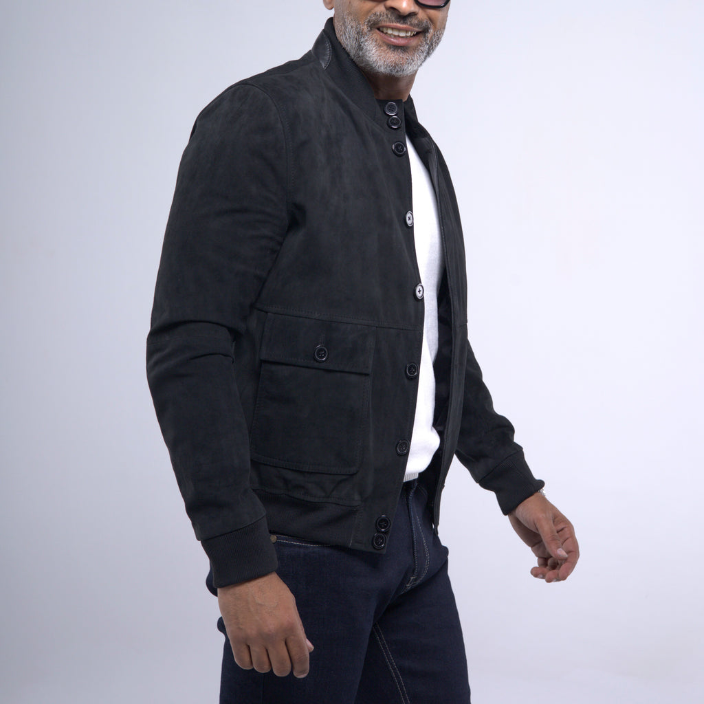 A man wearing a black suede jacket over a white shirt and dark jeans, photographed against a light gray studio background.
