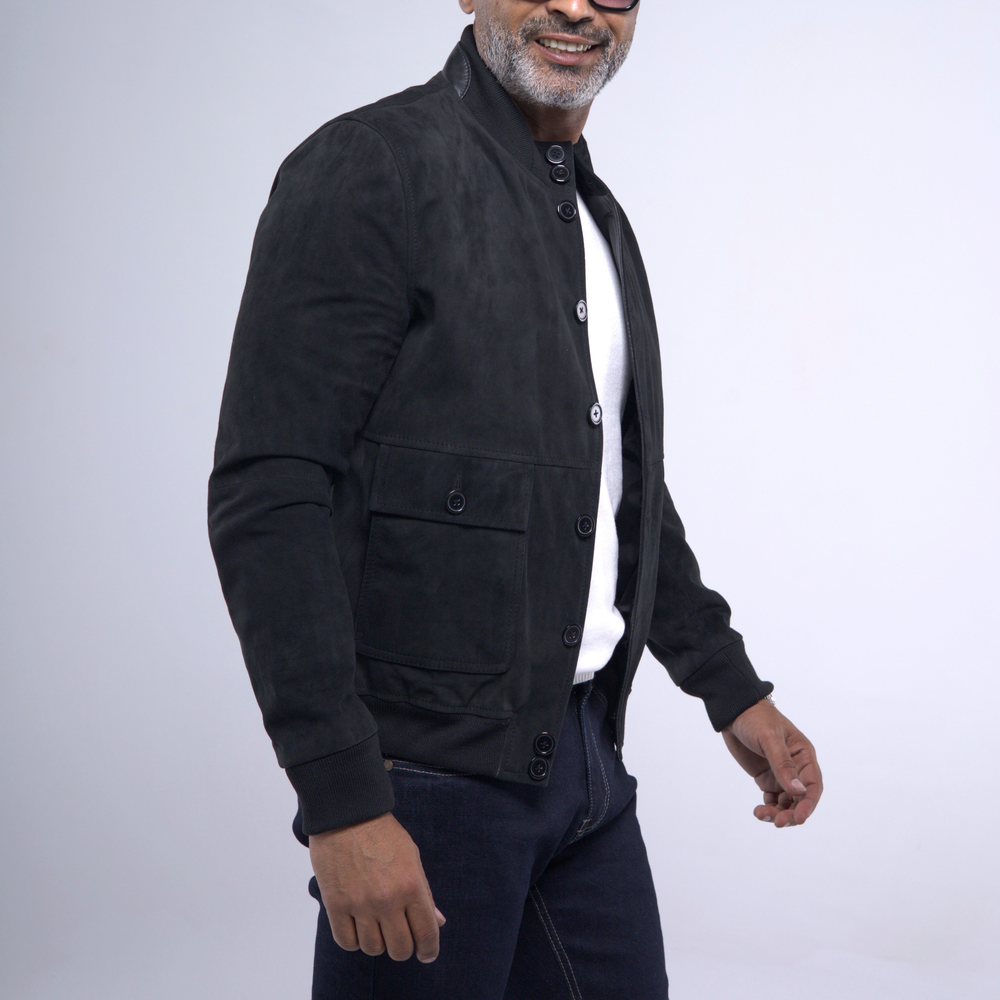 A man wearing a black suede jacket over a white shirt and dark jeans, photographed against a light gray studio background.