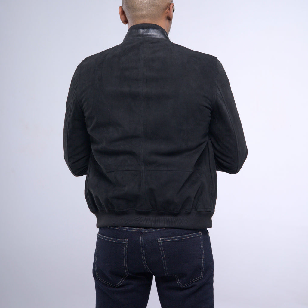 A man viewed from behind wearing a black suede or nubuck bomber jacket with a high collar and dark blue jeans, set against a plain light gray studio background.