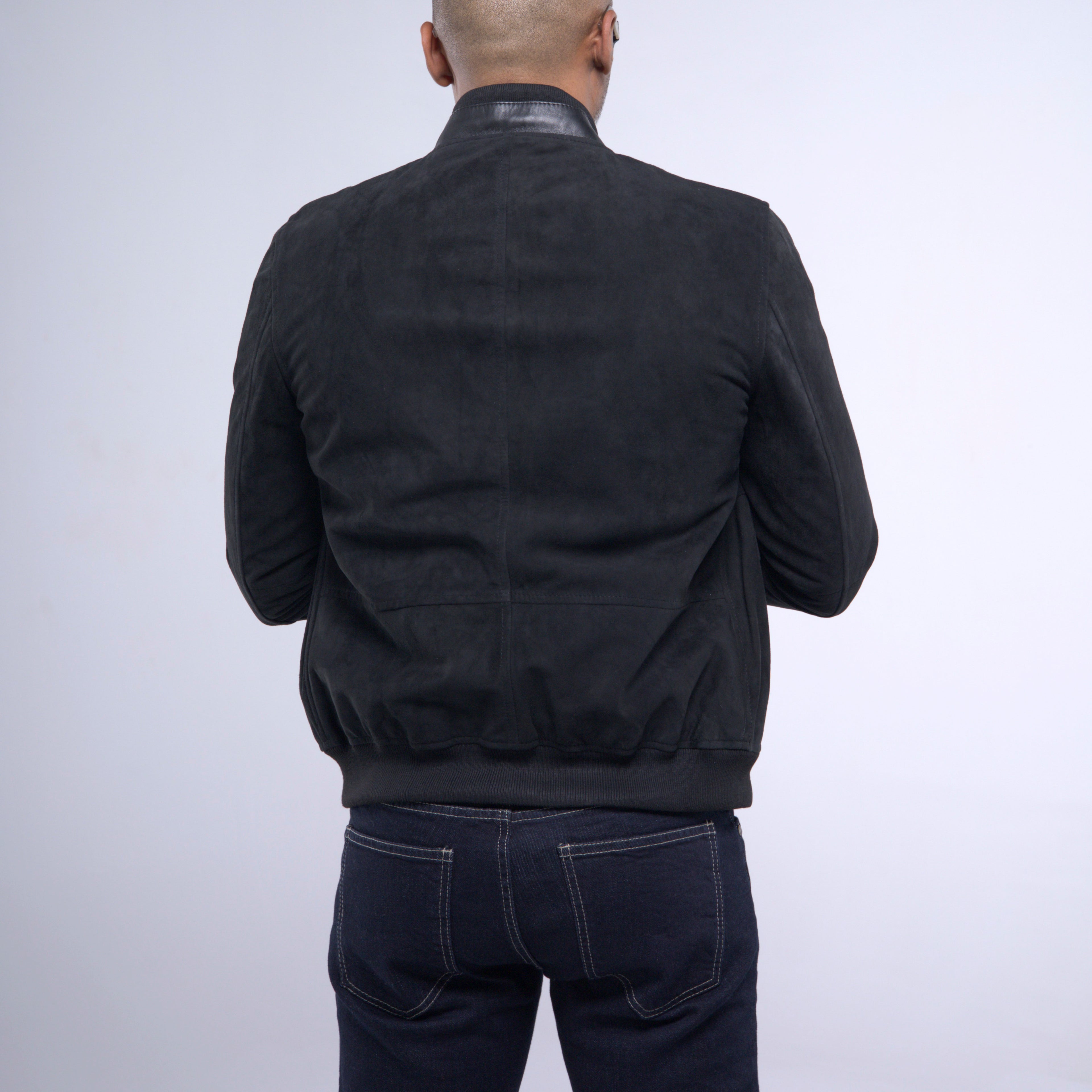 A man viewed from behind wearing a black suede or nubuck bomber jacket with a high collar and dark blue jeans, set against a plain light gray studio background.
