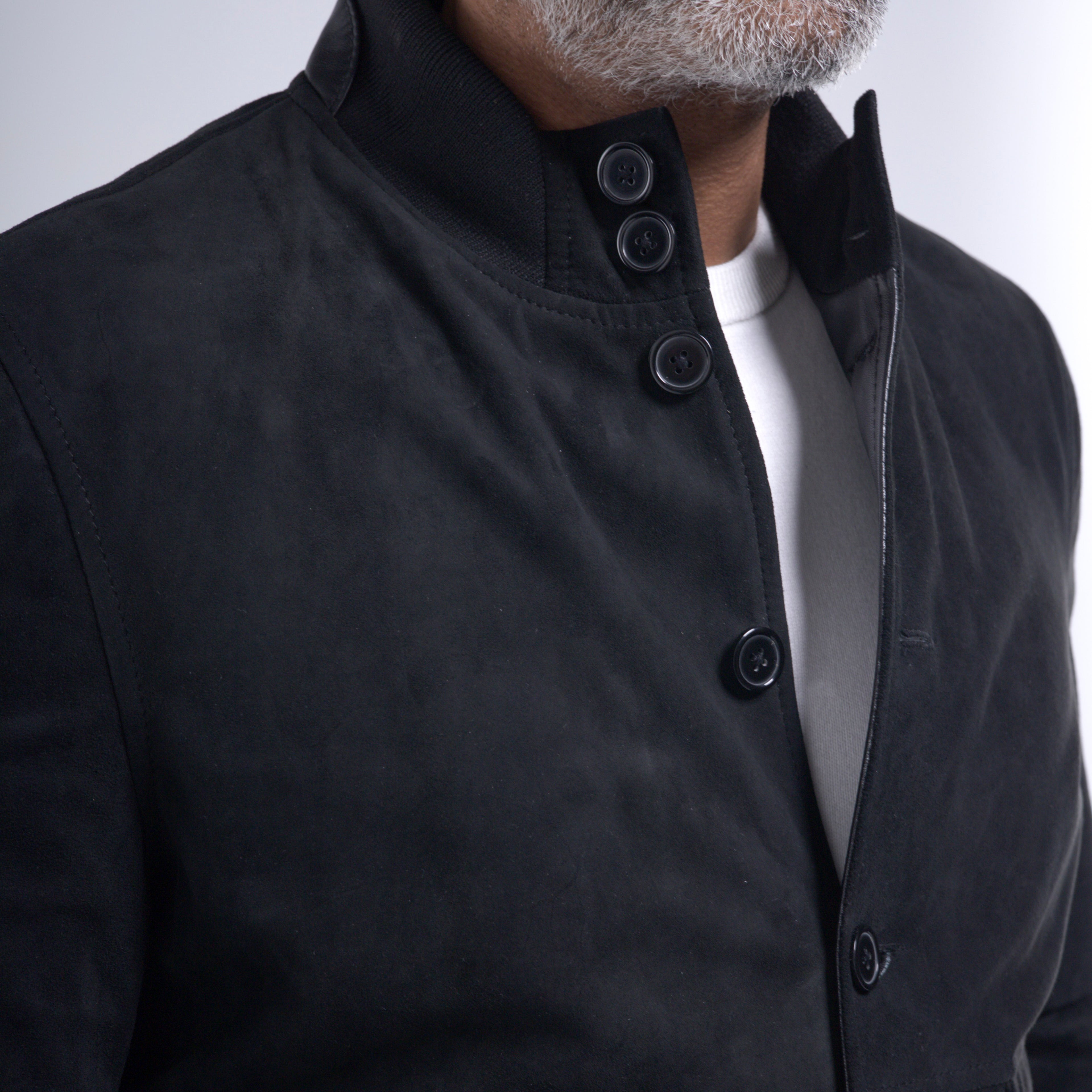 Close-up of a man wearing a black suede jacket with a high stand collar and black buttons, layered over a white t-shirt.