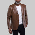 A man wearing a brown faux-leather blazer over a white t-shirt and black jeans, standing against a light gray studio background.