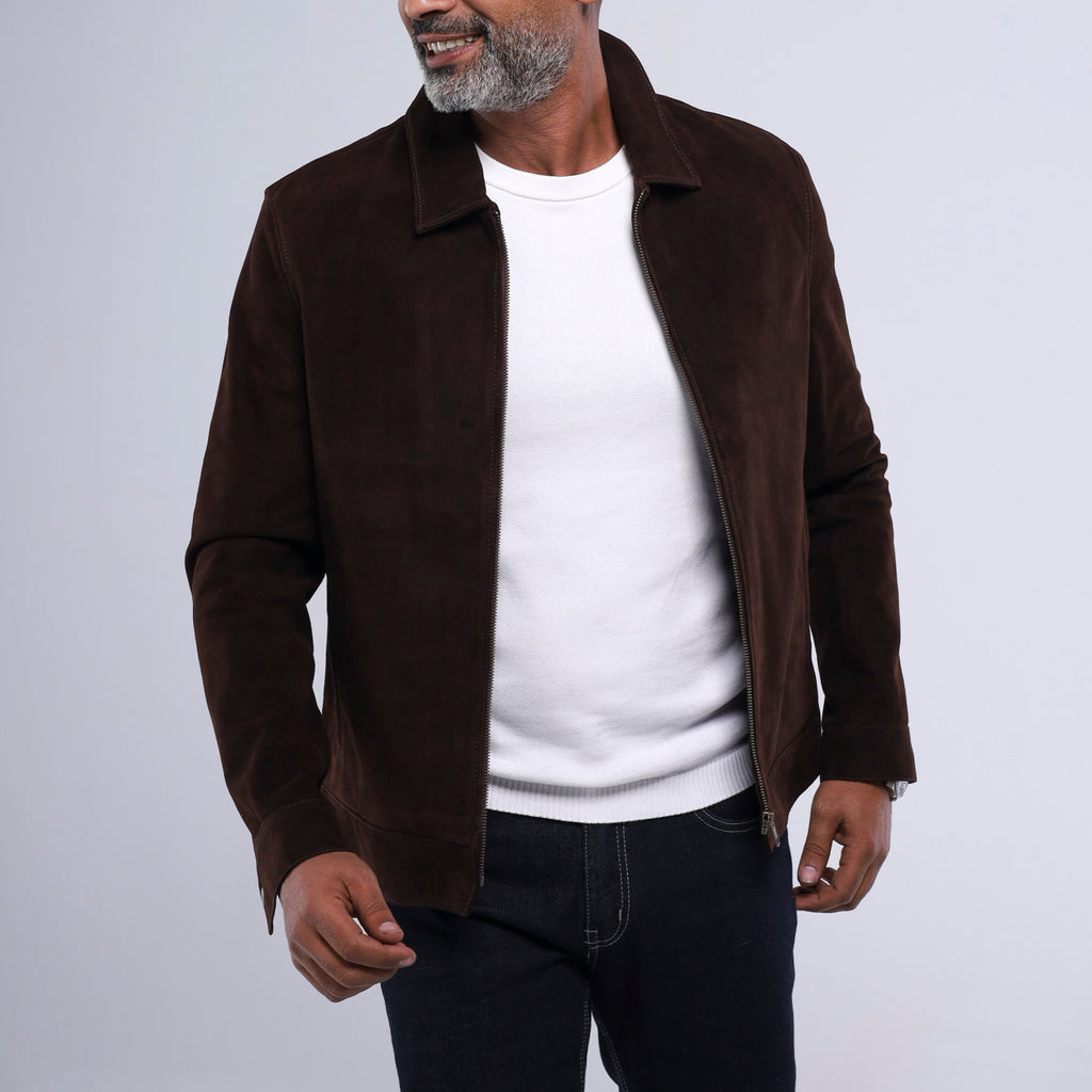 A man wearing a dark brown suede jacket over a white crewneck sweater and dark jeans, shown from the chin down against a light gray studio background.