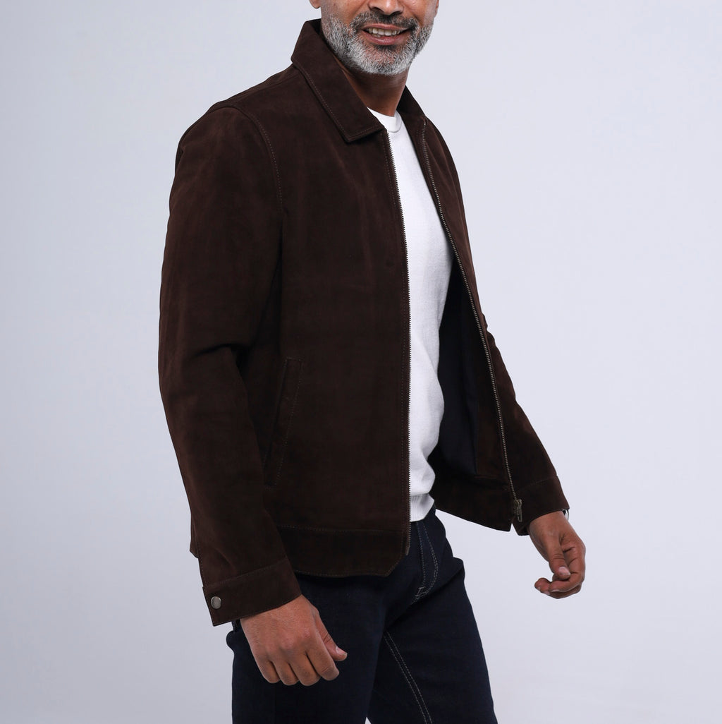 A man wearing a dark brown suede jacket over a white t-shirt and dark jeans, shown from the waist up against a light gray studio background.