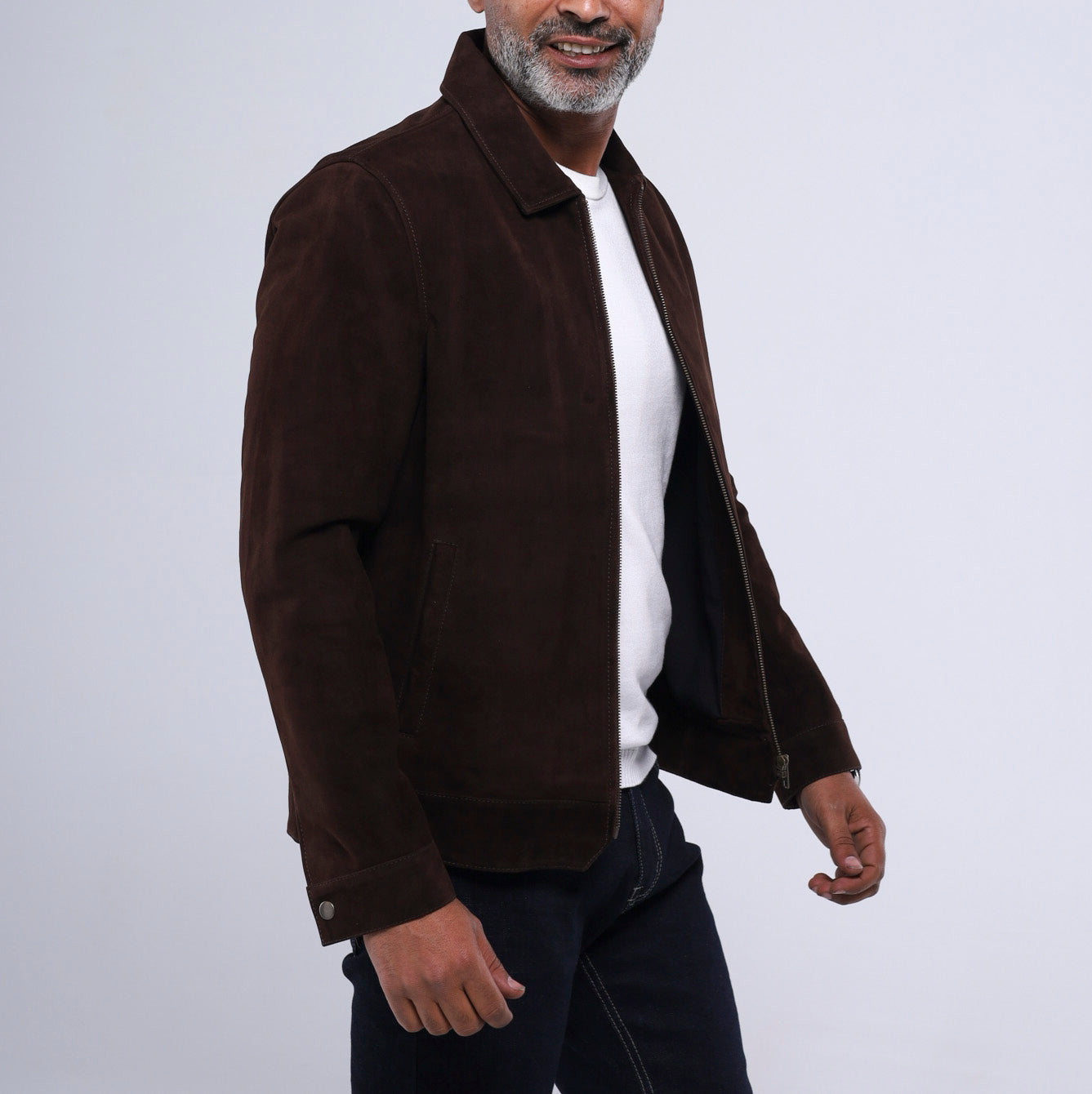 A man wearing a dark brown suede jacket over a white t-shirt and dark jeans, shown from the waist up against a light gray studio background.
