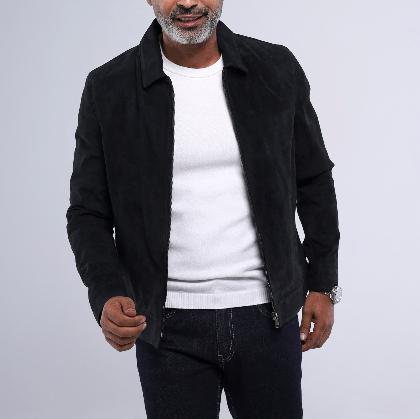 A man wearing a black suede jacket over a white crewneck sweater and dark jeans, photographed against a light gray studio background.