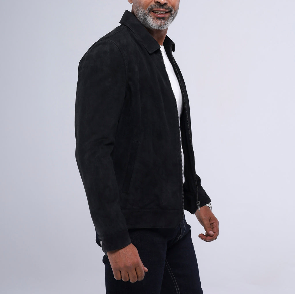 A man wearing a black suede jacket over a white t-shirt and dark jeans, shown from the waist up against a light gray studio background.