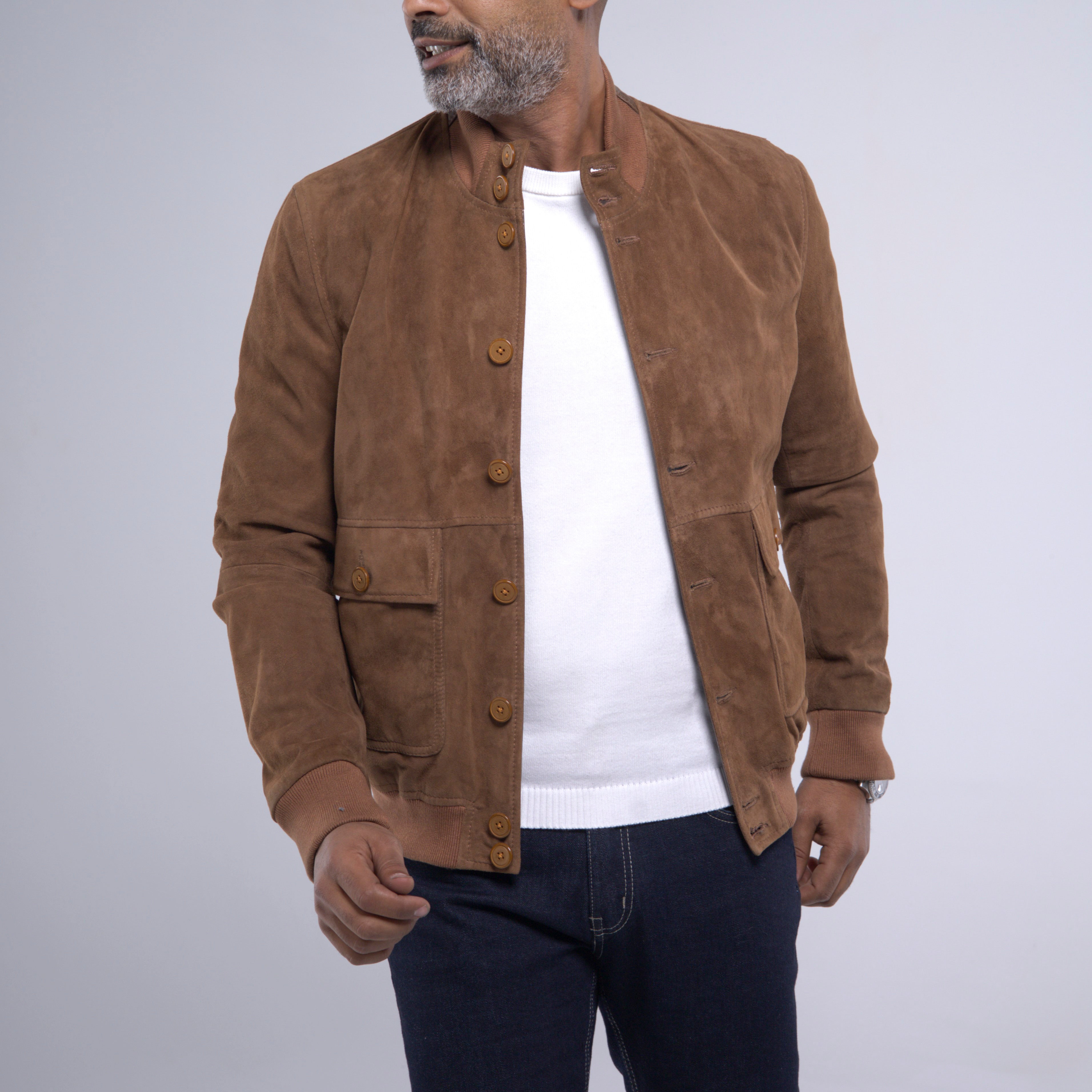 A man wearing a brown suede jacket over a white t-shirt and dark blue jeans, shown from the chin down against a light gray studio background.