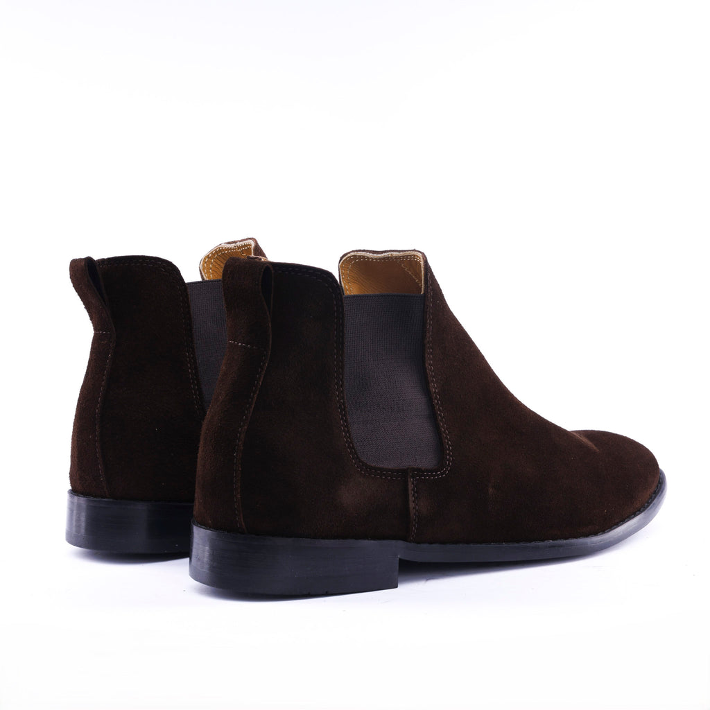 A pair of dark brown suede Chelsea boots with black elastic side panels and black soles, shown from a rear three-quarter angle on a white background.
