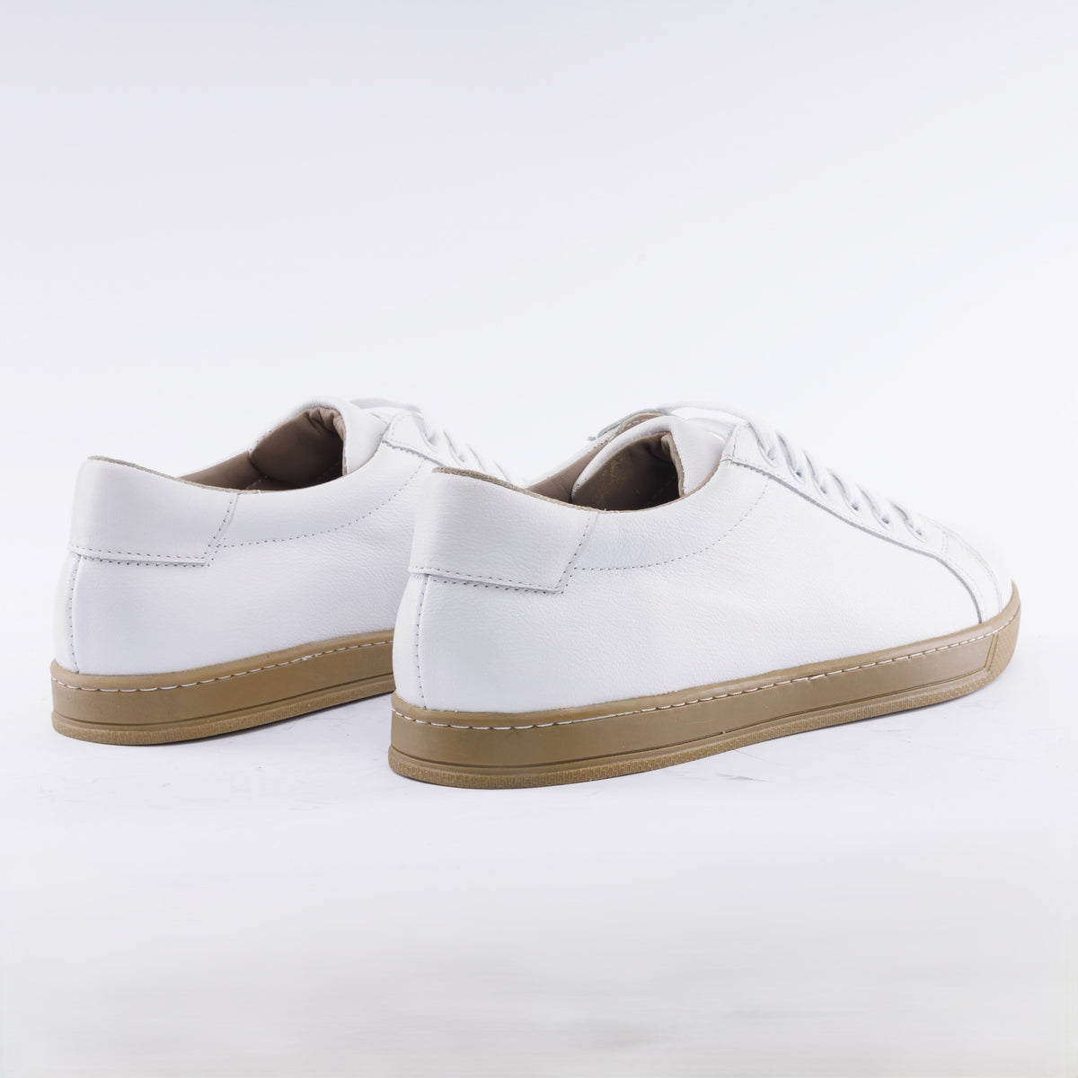 A pair of white leather low-top sneakers with white laces and tan rubber soles, shown from a three-quarters angle on a white studio background.