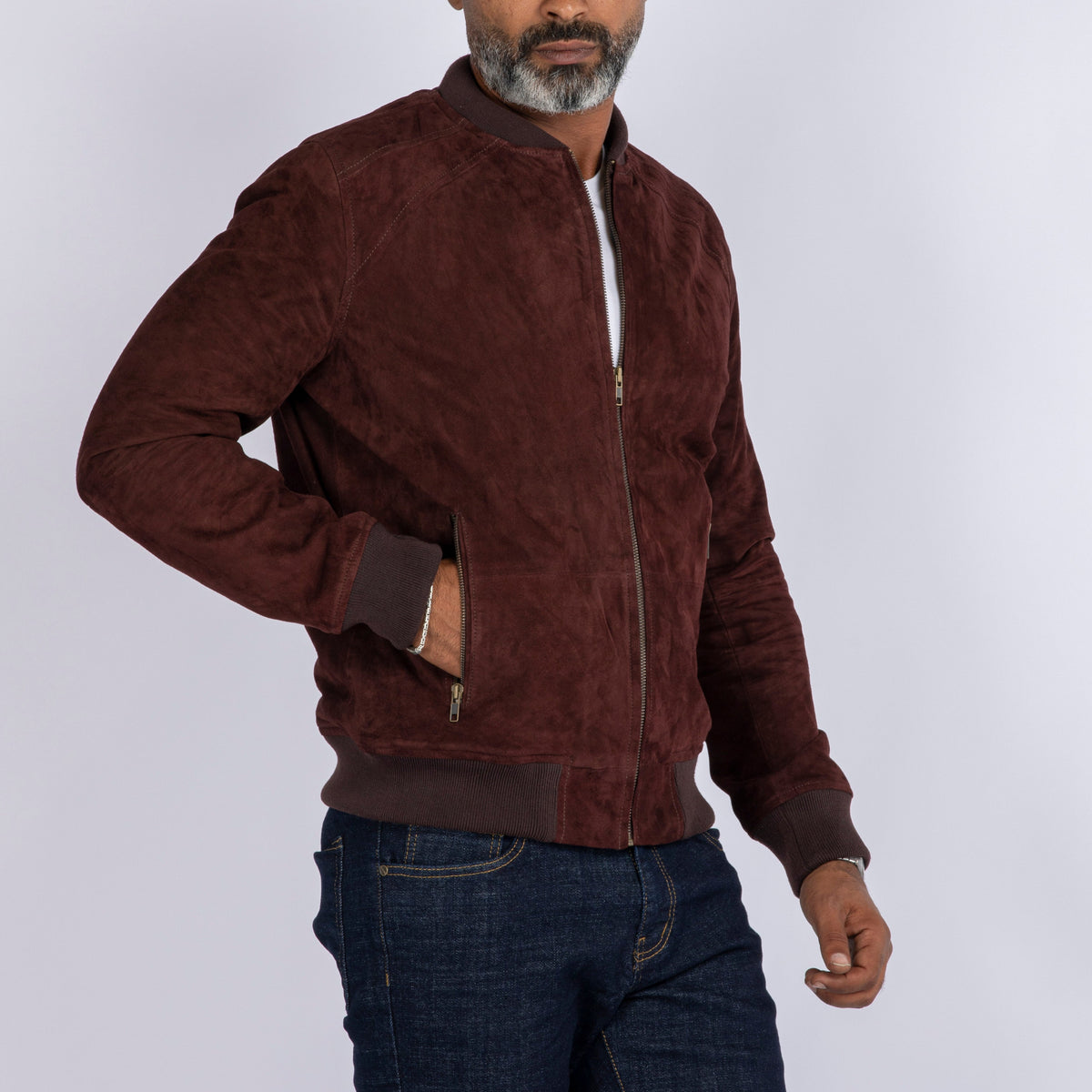 A man wearing a dark brown suede bomber jacket with a front zipper, ribbed collar, cuffs, and hem, paired with blue jeans, against a light gray studio background.