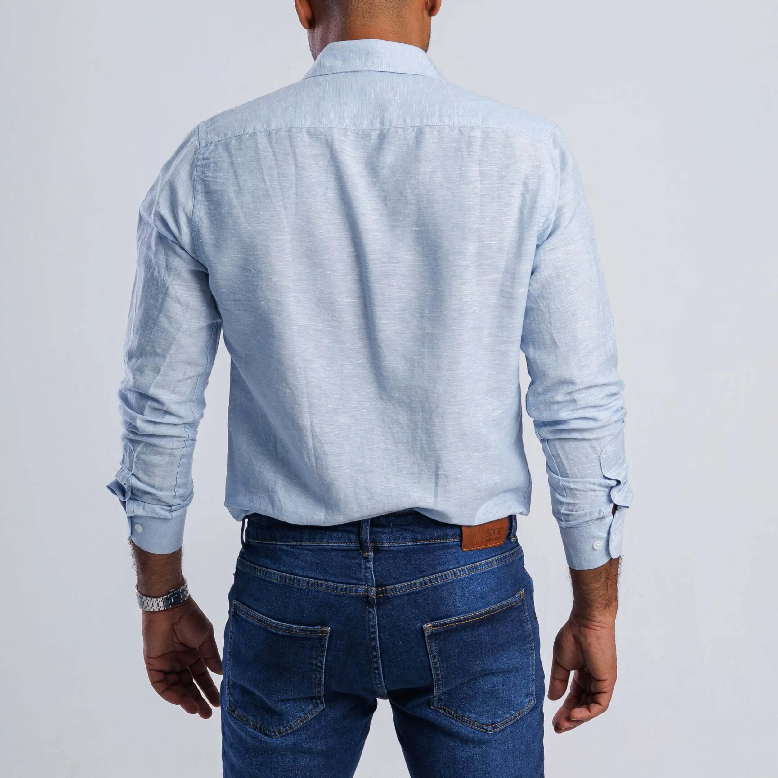 Back view of a man wearing a light blue linen blend slim fit shirt with long sleeves and casual jeans.
