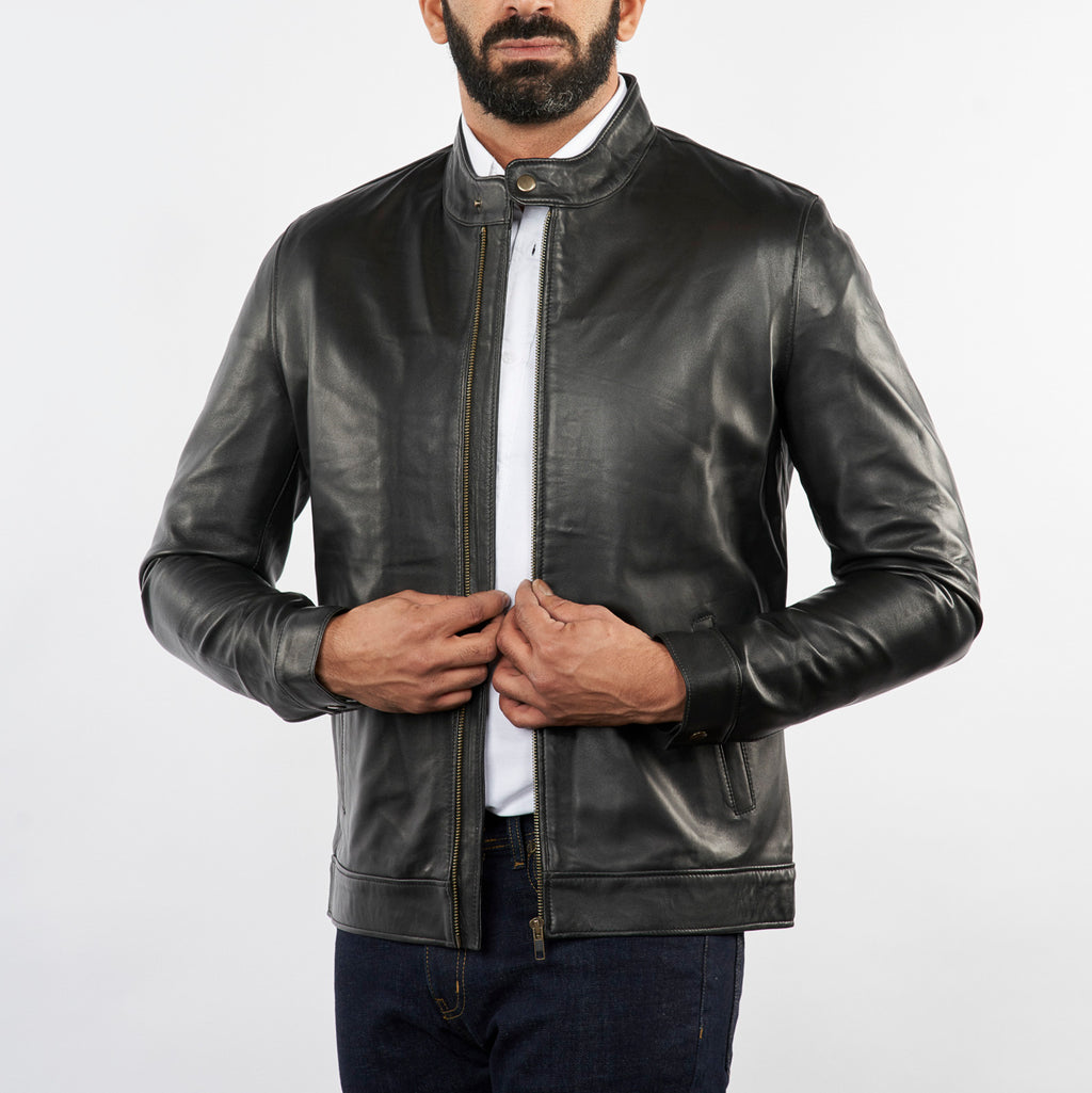 A man wearing a black leather jacket over a white collared shirt and dark jeans, standing against a light gray studio background.